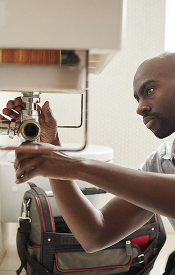 Young black male plumber sitting on the floor fixing a bathroom sink, seen from doorway